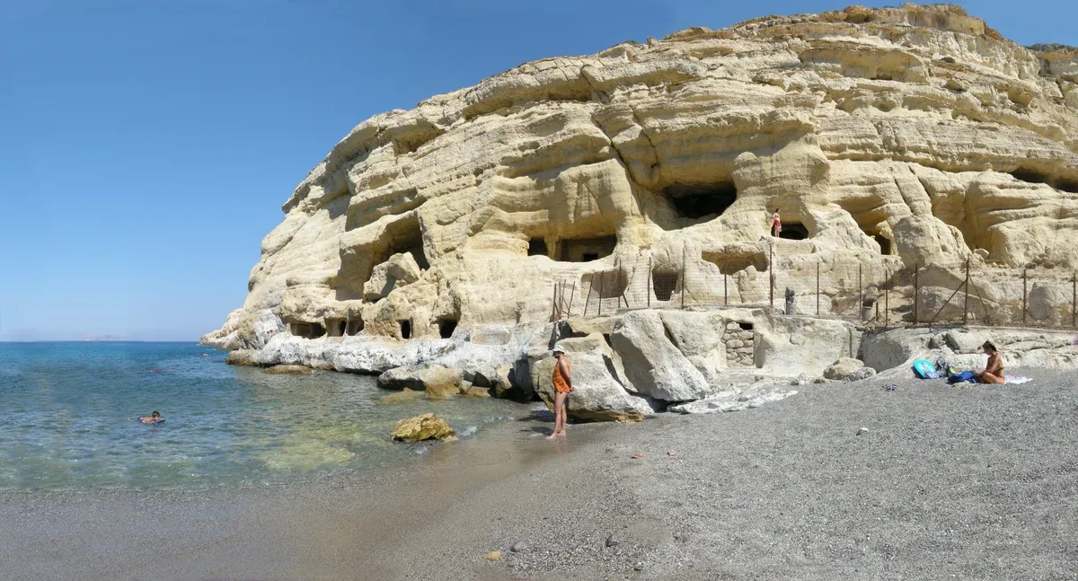 Swimmers in the clear water beneath the rock-cut caves of Matala beach, Crete